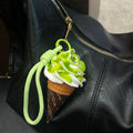 A realistic-looking ice cream cone charm with green matcha-flavored swirls and white cream, topped with small, shiny embellishments. It is attached to a light green braided rope with a metal clasp, hanging from the zipper of a black leather bag.