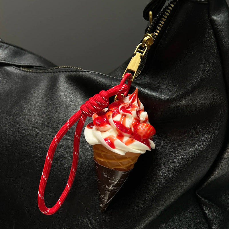 A realistic-looking ice cream cone charm with red strawberry-flavored swirls and white cream, topped with a small, detailed strawberry. It is attached to a red braided rope with a metal clasp, hanging from the zipper of a black leather bag.