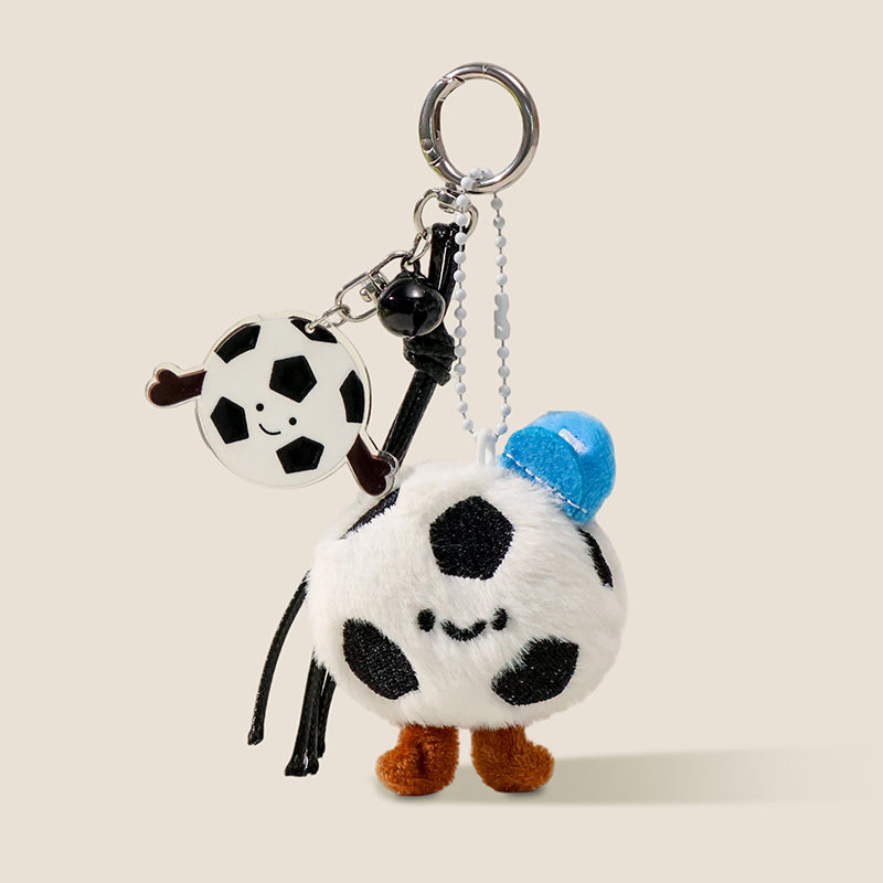 A plush soccer ball keychain with a smiling face, brown feet, and a blue hat, attached to a silver keyring with a matching acrylic soccer ball charm and a black bell.