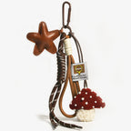 A leather star-shaped bag charm in brown, attached to a woven rope strap with brown and beige accents. A plush red mushroom with white pearl-like dots and a white fuzzy base hangs below, accompanied by a small fabric tag with a logo and text, designed as a cute and trendy accessory for bags or keychains.