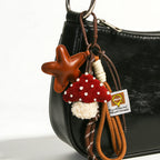 A leather star-shaped bag charm in brown, hanging from the handle of a black leather bag. The charm is attached via a woven rope strap with brown and beige accents, and a plush red mushroom with white pearl-like dots and a white fuzzy base is visible below, adding a playful touch to the bag.