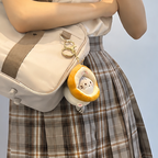 A person wearing a plaid skirt holds a beige shoulder bag with a taiyaki cat plush bag charm attached. The charm features a cute cat face peeking out from the taiyaki-shaped costume, adding a playful touch to the bag.