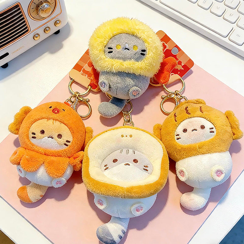 A collection of four different plush cat keychains in various food-themed hoods (orange, yellow, white, and brown) arranged on a pink surface. Each cat has a unique expression and color scheme, attached to gold-colored keyrings.