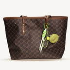A brown tote bag with a repeating 'MK' monogram pattern features a green and white tennis-themed keychain hanging from its strap, including a plush tennis ball, a miniature racket, and a rope handle.