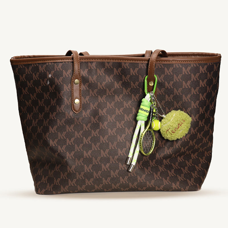 A brown tote bag with a repeating 'MK' monogram pattern features a green and white tennis-themed keychain hanging from its strap, including a plush tennis ball, a miniature racket, and a rope handle.