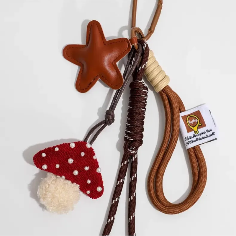 A brown leather star-shaped charm and a red knitted mushroom with white pearl-like dots and a white pom-pom base, attached to a brown carabiner with woven cords. A white tag with a logo is also attached.