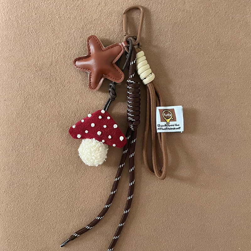 A brown leather star-shaped charm and a red knitted mushroom with white pearl-like dots and a white pom-pom base hanging from a brown tote bag. The charm is attached with woven cords and a brown carabiner, featuring a white logo tag.