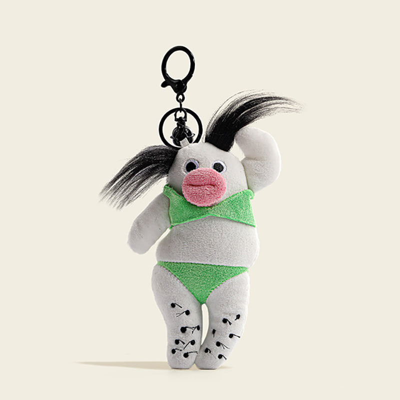 A plush bag charm in white fabric with green clothing, a pink mouth, and black pigtails. It is attached to a black carabiner clip with a small bell, featuring black dots on its body for a whimsical and trendy design.