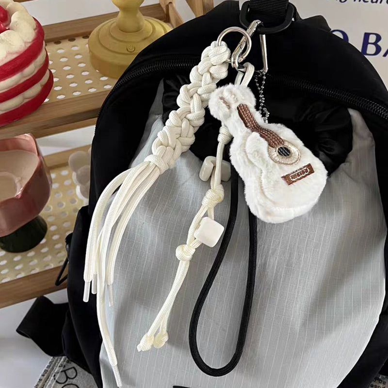 A close-up of a white plush guitar keychain with brown details, hanging from a black backpack with a white braided rope strap and white plastic beads.