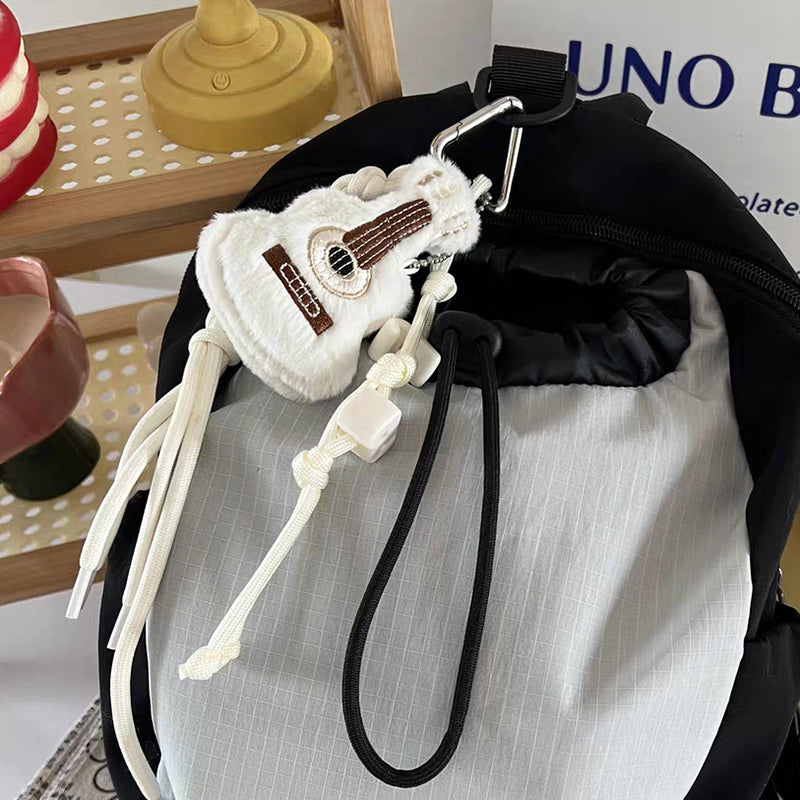 A white plush guitar keychain with brown details hanging from the zipper of a black and grey backpack, featuring a white braided rope strap.