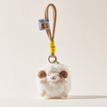 A front-facing view of a white fluffy sheep plush keychain with brown spiral horns and a smiling face, attached to a beige cord with a yellow wrap and a small branded tag.