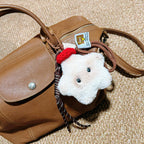 A close-up of a white plush star-shaped doll with black button eyes and a red knitted hat, hanging from a brown leather bag with a woven strap and a small tag.