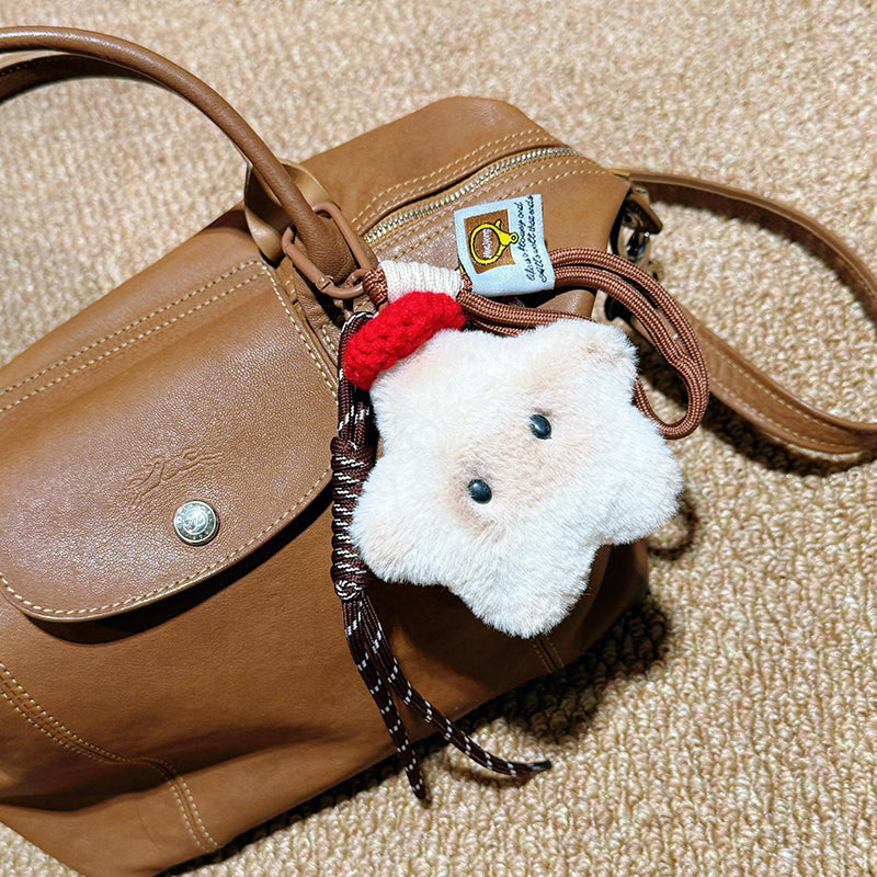 A close-up of a white plush star-shaped doll with black button eyes and a red knitted hat, hanging from a brown leather bag with a woven strap and a small tag.