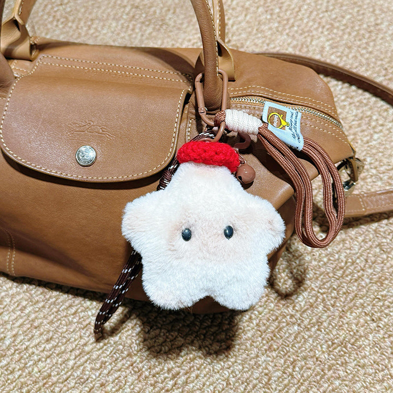 A white plush star-shaped doll with black button eyes and a red knitted hat, attached to a brown leather handbag with a woven strap and a small tag.