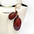 A white pebbled leather handbag accessorized with a brown leather coffee bean pendant. The charm hangs from the side handle, adding a rustic touch to the modern bag design.