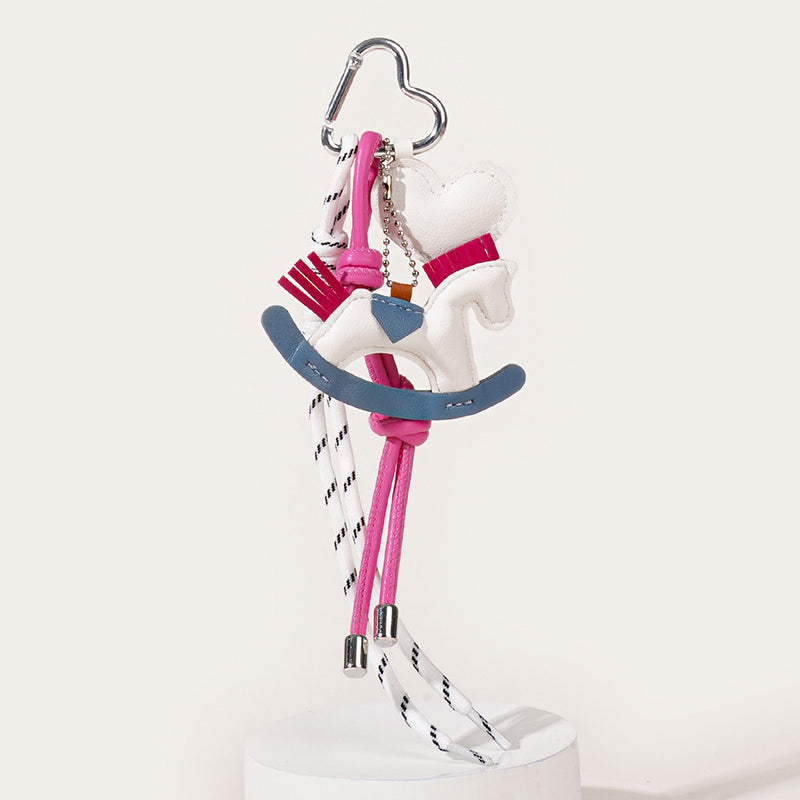 A stylish bag charm displaying a white leather rocking horse with pink accents and a blue base. It hangs from a silver clasp with white patterned ropes and solid pink cords.
