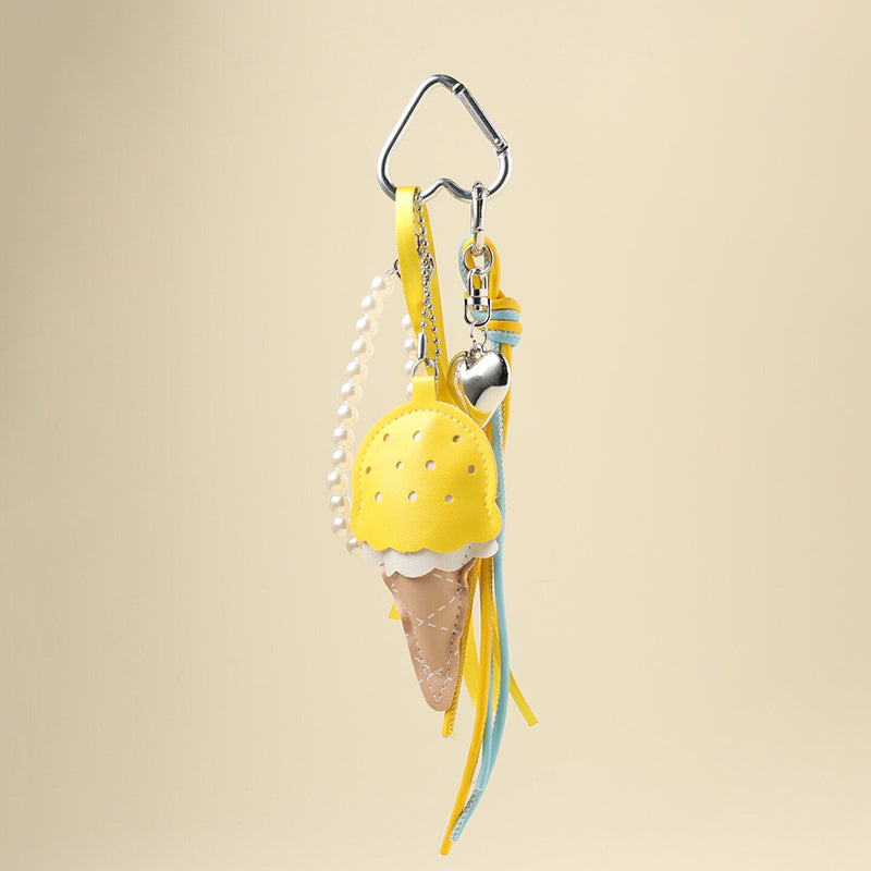 A yellow leather ice cream cone bag charm with a white scalloped edge and perforated details, attached to a silver carabiner clip. It features yellow and light blue ribbons, a pearl chain, and a small silver heart charm, designed as a cute and trendy accessory for bags or keychains.