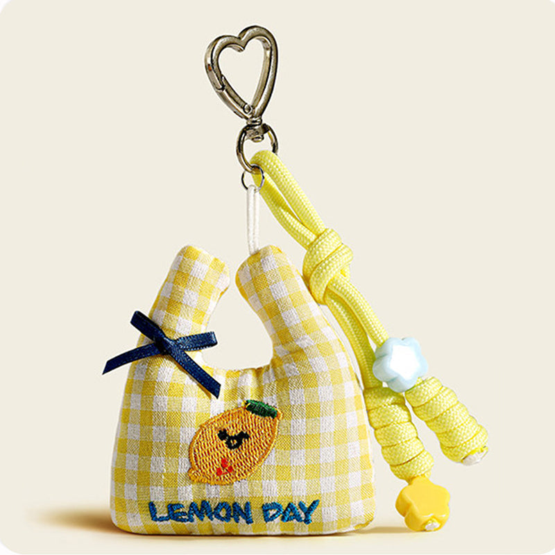 A yellow and white plaid fabric bag charm shaped like a mini tote, with an embroidered lemon and "LEMON DAY" text, decorated with a blue bow and yellow flower beads.
