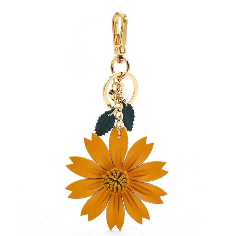 A hanging view of a handcrafted leather keychain shaped like a yellow daisy. It features a gold-tone metal clasp and ring, with two dark green leaf charms attached to the chain.
